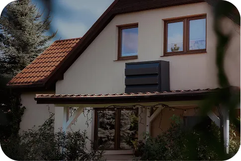 A detached house with a beige façade and orange roof. Dark Silencions s|AC| acoustic enclosures are mounted on the wall of the building, effectively silencing the air conditioner and improving the aesthetics.