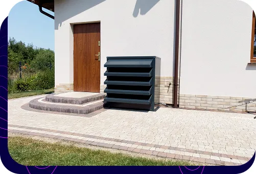 A modern, graphite-coloured acoustic enclosure for a heat pump, aesthetically integrated into the surroundings of a detached house at the entrance.