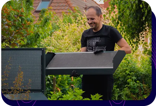 A man in a garden, installing the upper part of the dark Silencions s|AC| acoustic enclosure, smiling and demonstrating how easy it is to install.