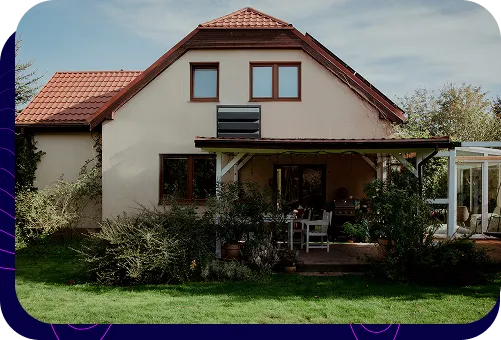 A detached house with a beige façade and brown roof. A dark Silencions s|AC| acoustic enclosure is mounted on the wall of the building to protect the outdoor unit of the air conditioner.