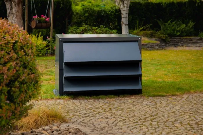Dark grey soundproof enclosure for a heat pump surrounded by greenery. Protects the external unit of the heat pump.