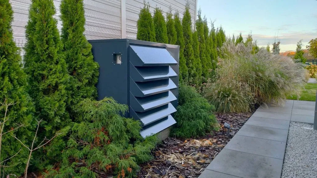 A modern, dark grey custom-made enclosure for a water chiller, aesthetically integrated into a well-kept garden with a row of thuja trees.
