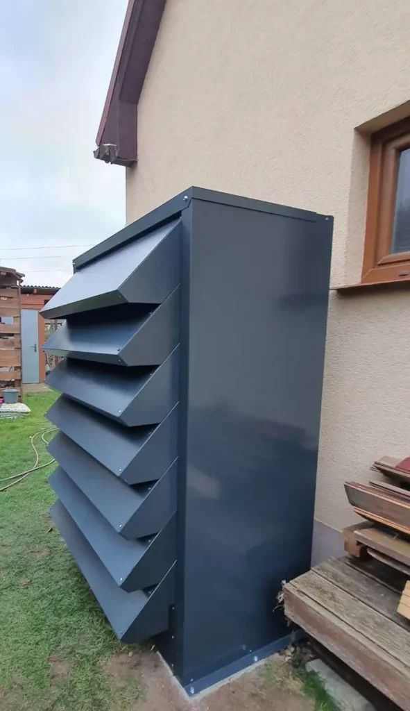 A tall, customised anthracite-coloured heat pump enclosure mounted on the wall of the house.