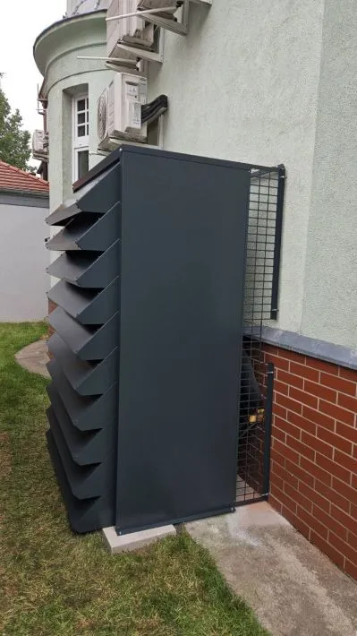 A tall, anthracite-coloured custom-made enclosure protecting a large heat pump, mounted on the wall of the building.