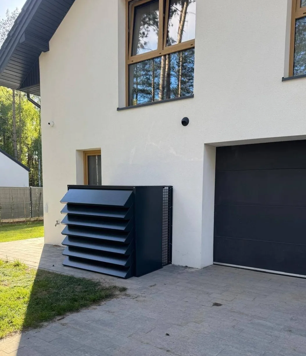 Dark grey, customised enclosure for the Silencions heat pump, perfectly matching the white façade and dark garage door of a modern house.