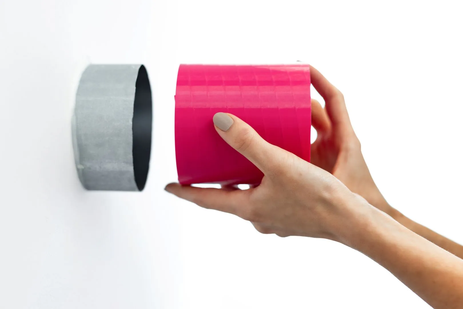 The photo shows hands inserting a pink s|TWIST sound attenuator into a round ventilation duct in a white wall. The scene demonstrates simple and clean installation, a perfect fit for the product, and easy integration into the ventilation system.
