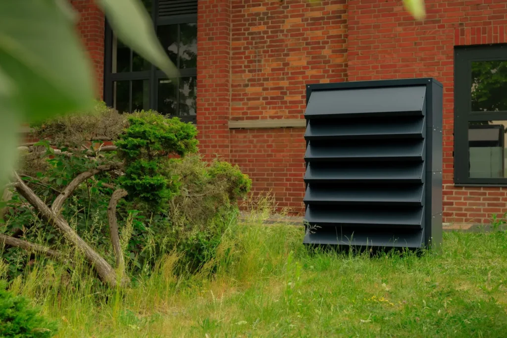 Modern, graphite-coloured soundproofing hood for an air conditioning unit on a brick building.