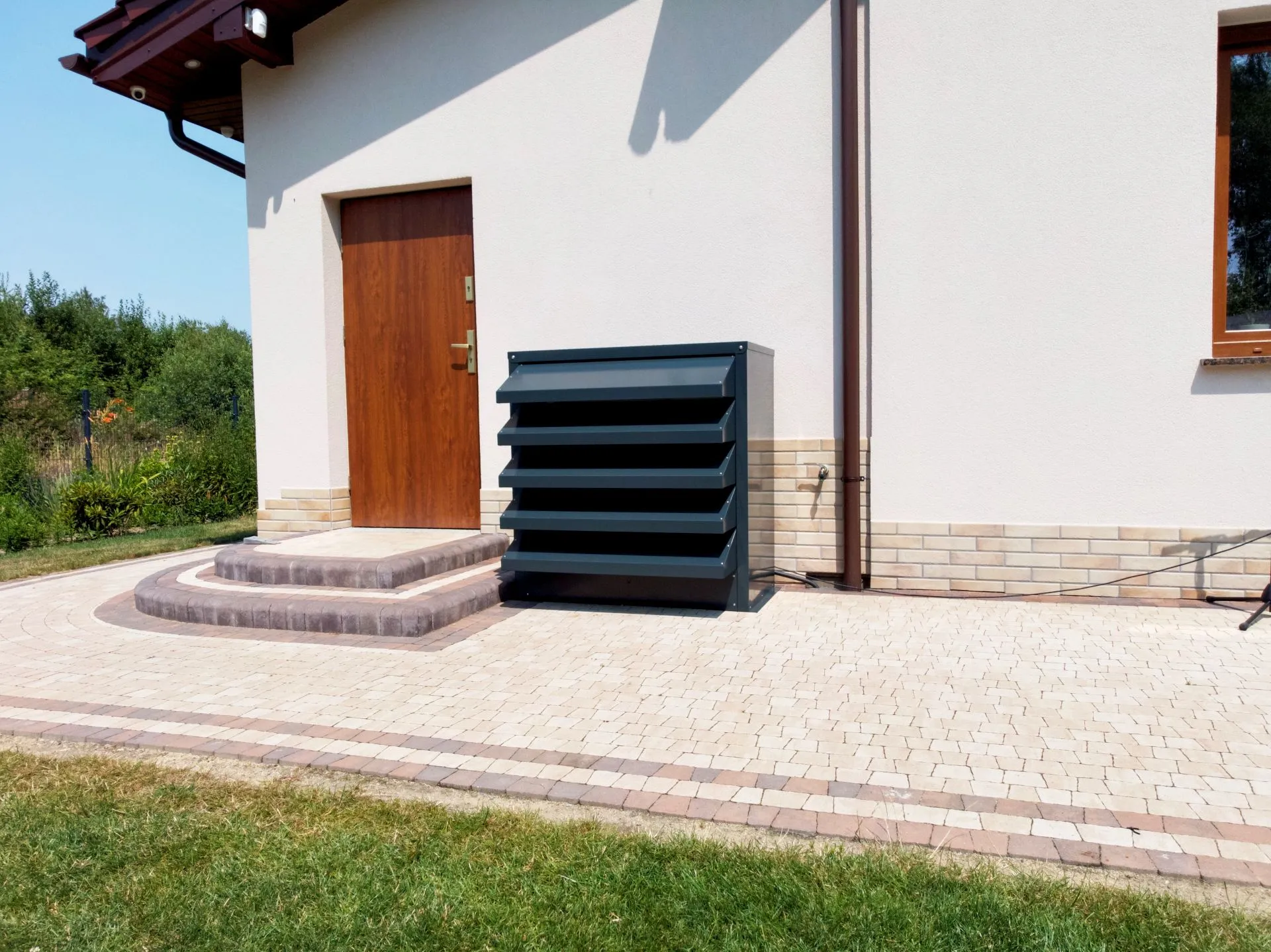 A modern, graphite-coloured acoustic enclosure for a heat pump, aesthetically integrated into the surroundings of a detached house at the entrance.