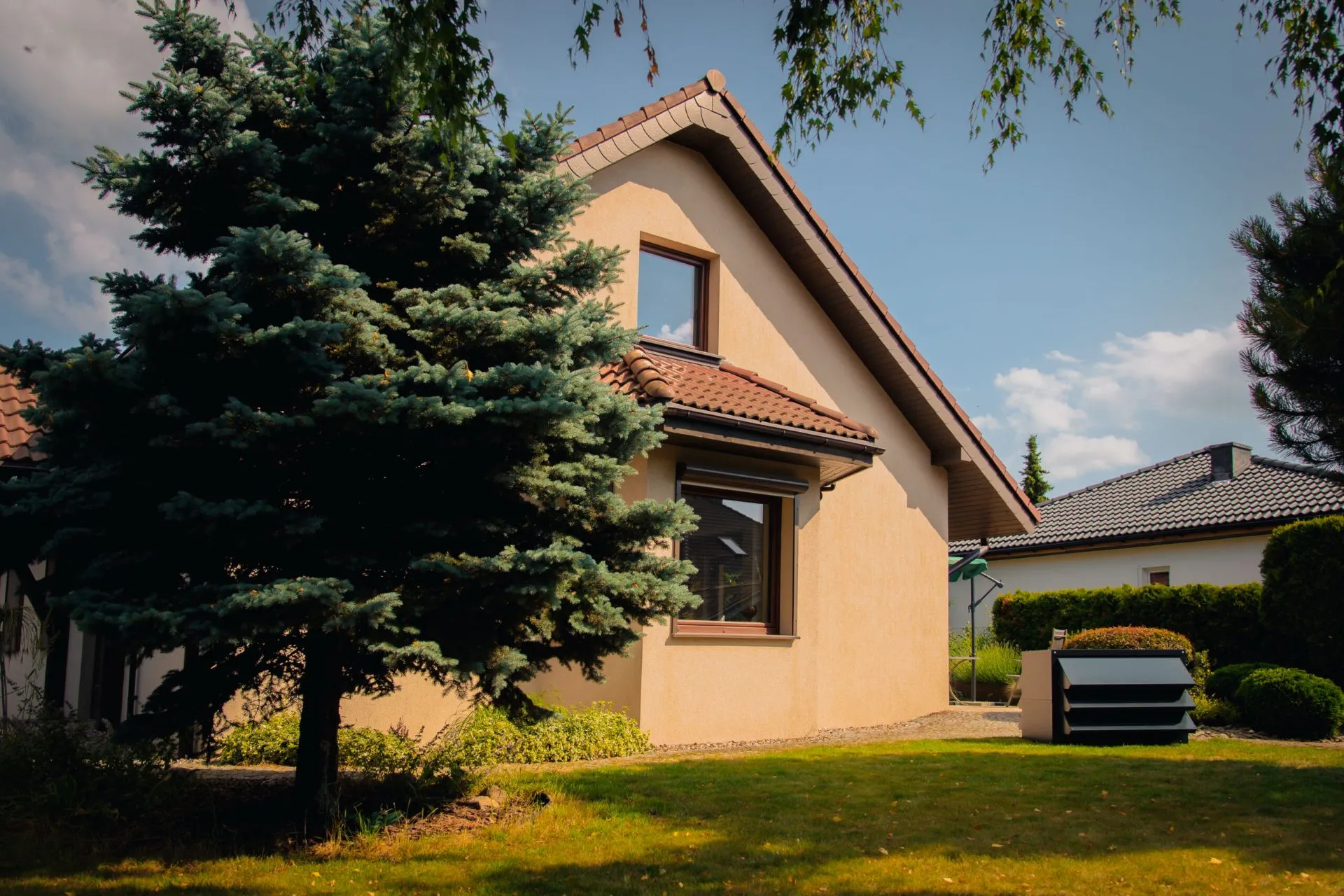 A detached house with a beige façade and an orange roof. Next to the house, on the lawn, there is a heat pump with a modern Silencions acoustic enclosure for heat pumps.