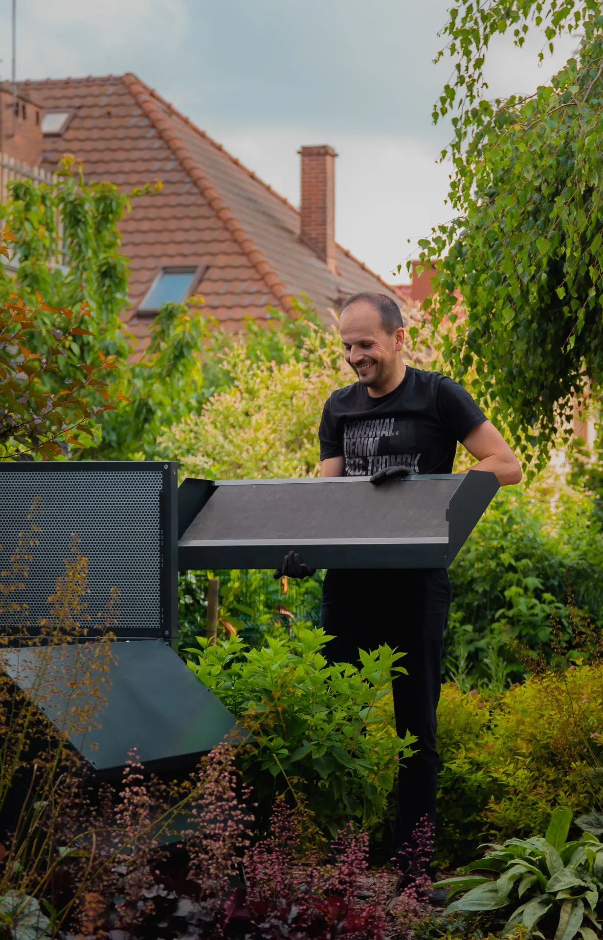 A man in a garden, installing the upper part of the dark Silencions s|AC| acoustic enclosure, smiling and demonstrating how easy it is to install.